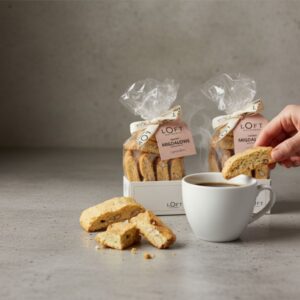 Biscotti-migdałowe-1-blog-Loft-Kulinarny Biscotti migdałowe Loft Kulinarny z kawą – dwie paczki ciastek na betonowym tle, dłoń podaje kromkę