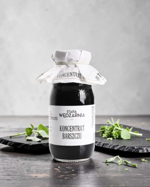Koncentrat barszczu czerwonego Stara Wędzarnia - na czarnym kamiennym talerzu, drewniany stol, szare cementowe tlo, zielone ziola
