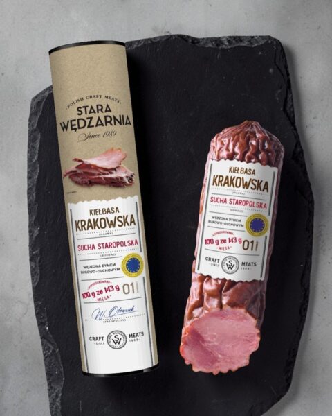 Krakowska sucha GTS w tubie Stara Wędzarnia - na czarnym kamiennym talerzu, drewniany stol, szare cementowe tlo, zielone ziola