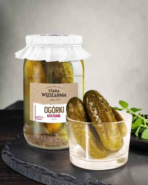Ogórki kiszone Stara Wędzarnia - na czarnym kamiennym talerzu, drewniany stol, szare cementowe tlo, zielone ziola